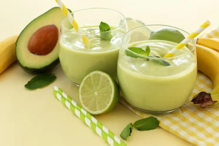 A glass filled with a creamy green smoothie made with avocado, spinach, banana, pineapple, and coconut water, surrounded by ingredients, representing the Avocado Spinach Delight Smoothie recipe.