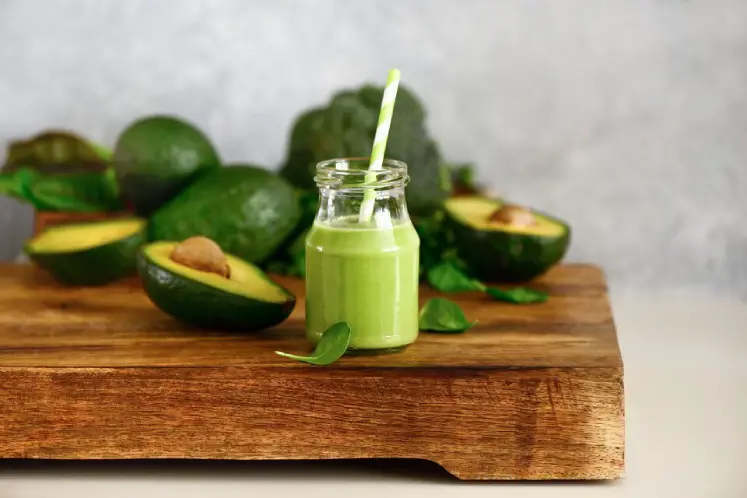 A glass filled with a vibrant green avocado spinach smoothie, garnished with a slice of avocado and spinach leaves, surrounded by fresh avocados and spinach, representing a nutritious smoothie recipe for healthy weight gain.