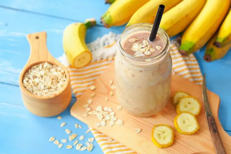 A glass filled with a creamy banana peanut butter smoothie topped with a slice of banana and a dollop of peanut butter, surrounded by fresh bananas and peanut butter jar, representing a delicious and nutritious smoothie recipe for healthy weight gain.