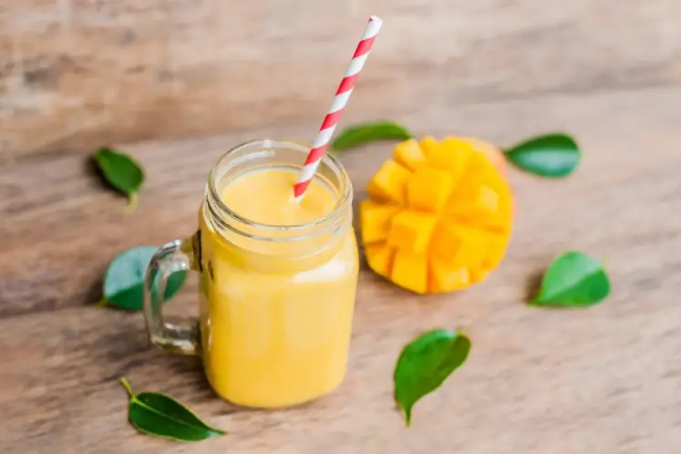 A glass filled with a refreshing yellow mango coconut smoothie, garnished with a slice of mango and coconut flakes, surrounded by fresh mangoes and coconut, representing a tropical and nutritious smoothie recipe for healthy weight gain.