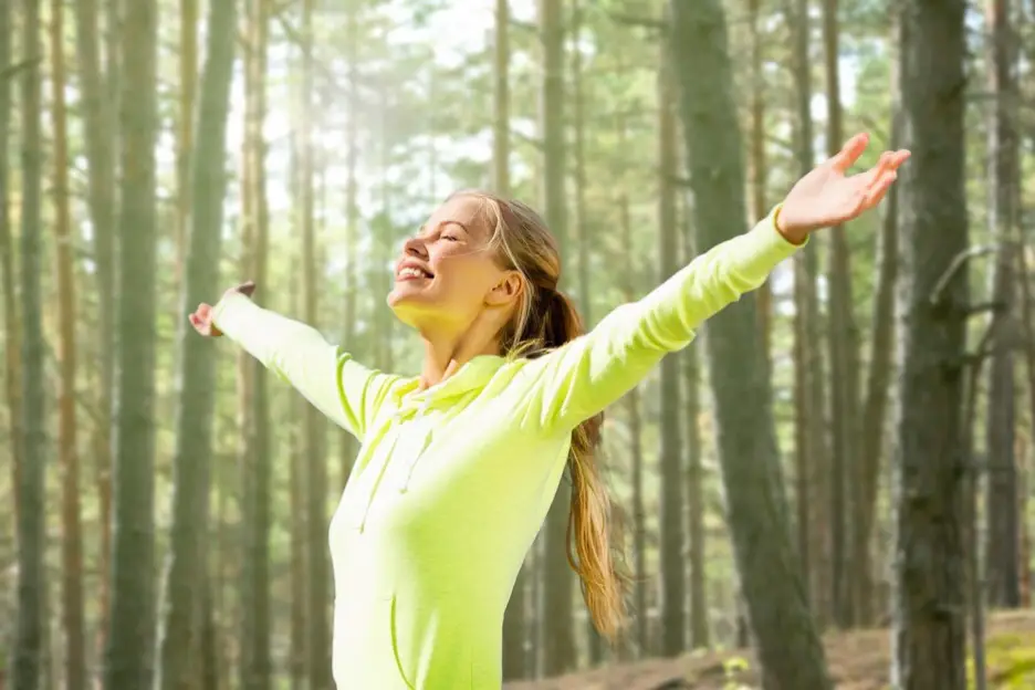 Happy woman raising hands over woods background, representing the positive impact of living healthier and adopting healthy lifestyle habits.