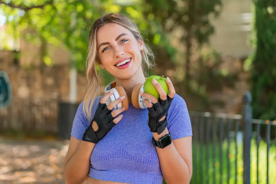 Alt Text: A smiling woman in workout attire holding a green apple and headphones, representing healthy lifestyle choices with exercise and nutrition.