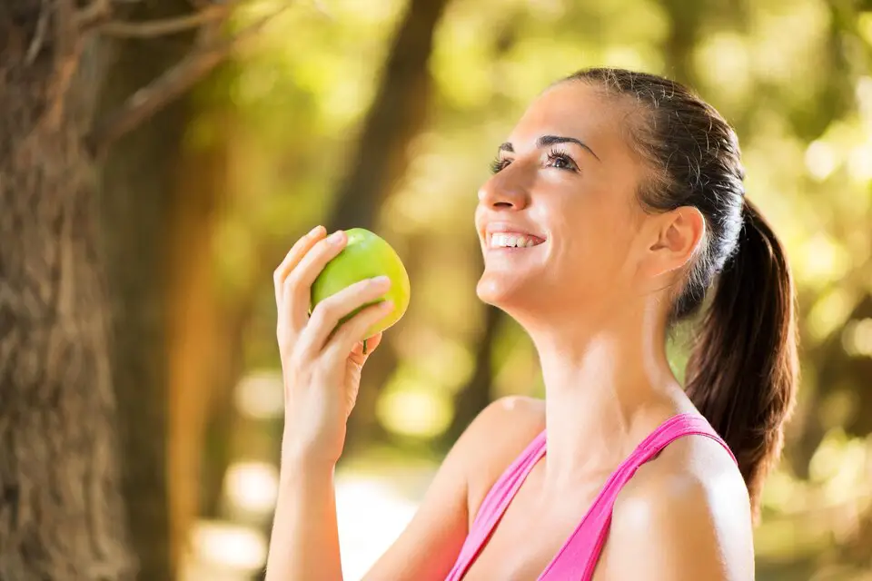 A smiling woman in a pink tank top holding an apple outdoors, representing a healthy lifestyle and balanced nutrition.