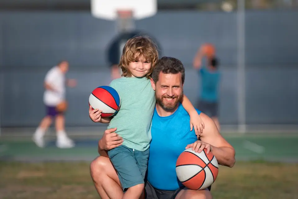 Father and young son playing basketball on an outdoor court, showcasing exercise routines for children aged 5-12.