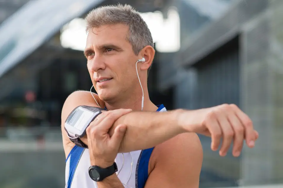 Middle-aged man wearing earphones and an armband stretching his arm before exercising, emphasizing fitness routines for adults aged 36-55.