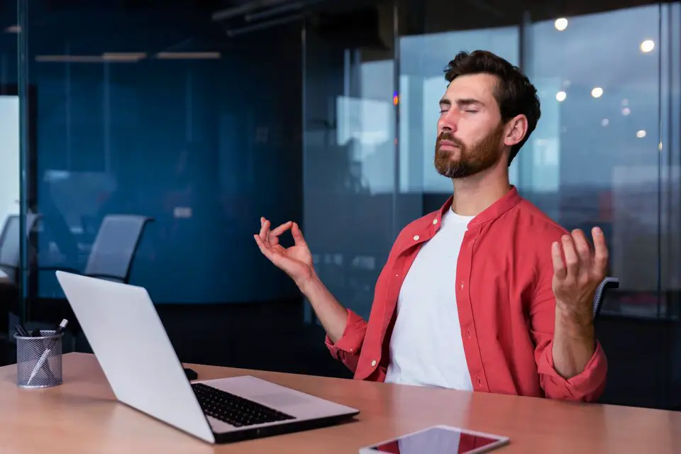 A man in a red shirt meditating at his desk in an office setting, representing the integration of health and wellness practices into a professional environment.