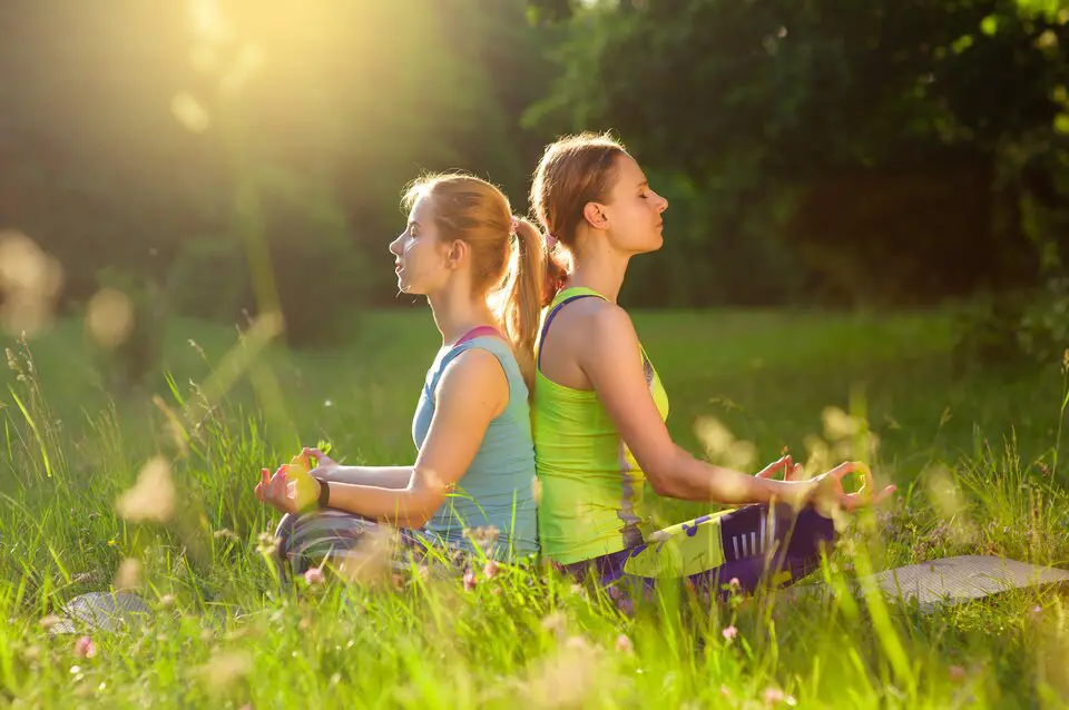 Two women sitting back-to-back, meditating in a sunlit grassy field, representing relaxation and mindfulness for improved health and wellness.