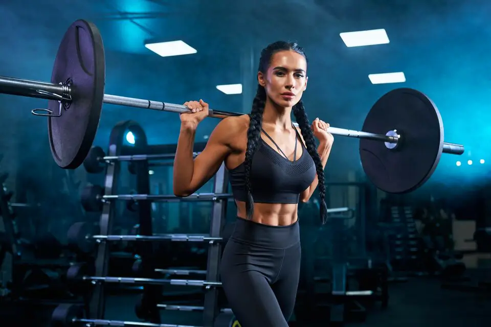 A fit woman with braided hair lifting a barbell on her shoulders in a gym, showcasing strength and dedication to fitness strategies.